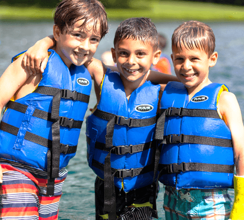 Three boys with life vests on smiling for a group photo in front of the lake