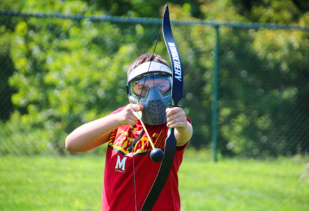 Camper with bow and arrow practicing archery with protective mask on