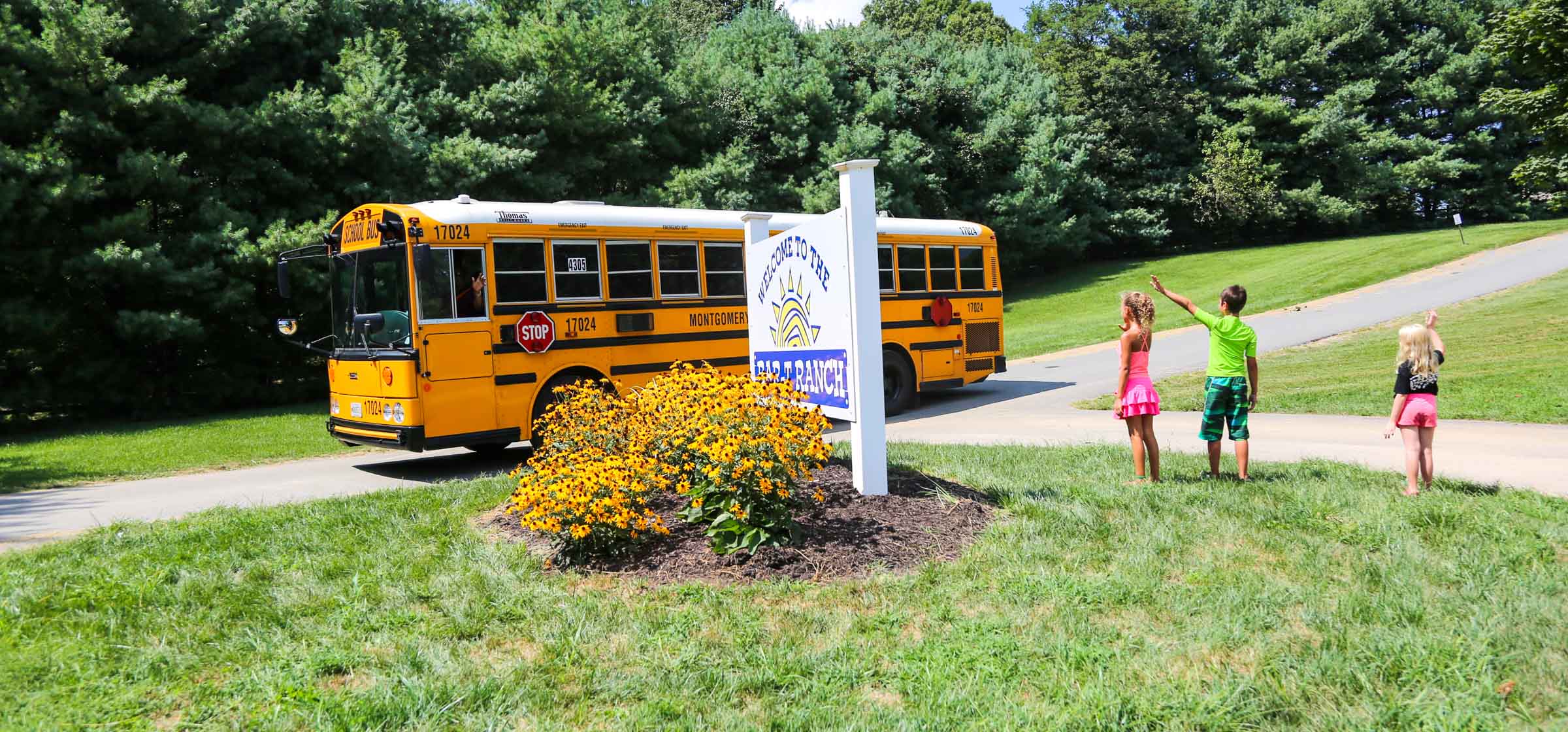 Campers on a grassy lawn waving to a yellow bus