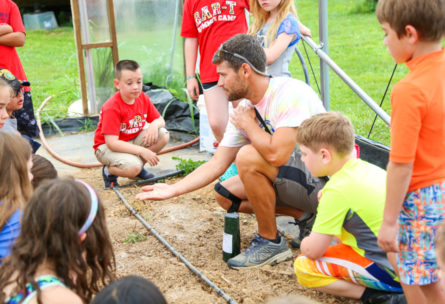 Teacher kneeling with seeds in his palm showing students gathered around in a greenhouse