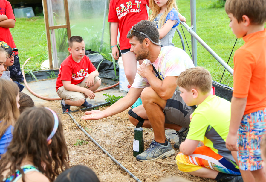 Teacher kneeling with seeds in his palm showing students gathered around in a greenhouse