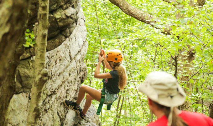 Girl camper with helmet and harness climbing up a rock outside in the woods