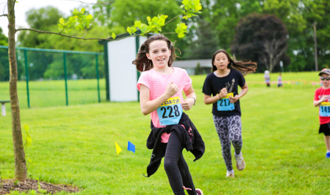 Two campers with numbers attached to their shirts running on a field in a 5K race
