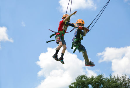 Two boys with helmets and gear swinging from a high beam with just the sky behind them